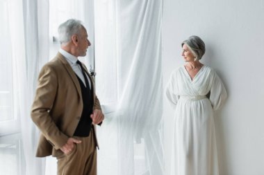 bearded middle aged man in suit posing while looking at cheerful bride in white dress near white curtains  clipart