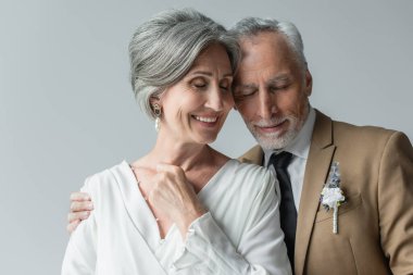 cheerful middle aged man in suit with floral boutonniere and woman in white wedding dress isolated on grey