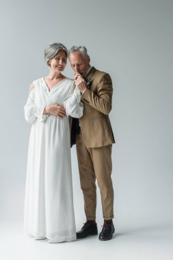 full length of middle aged man in suit kissing hand of happy bride in white wedding dress on grey