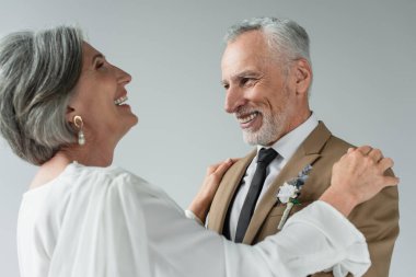 happy middle aged bride in white wedding dress hugging bearded groom and laughing isolated on grey