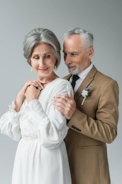 middle aged man in suit with floral boutonniere hugging smiling woman in white wedding dress isolated on grey
