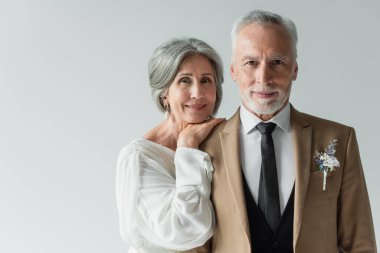 happy middle aged bride in white wedding dress leaning on shoulder of bearded groom isolated on grey