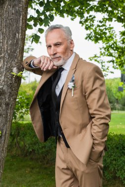 bearded middle aged groom in beige suit with boutonniere smiling in green park 