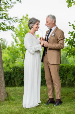 full length of happy middle aged groom in suit holding hands with mature bride in white dress in park