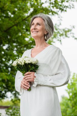 low angle view of happy middle aged bride in white dress holding wedding bouquet in green garden 