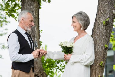side view of happy mature man in formal wear holding hand of bride with wedding bouquet in garden