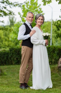 full length of pleased mature man in formal wear hugging bride with wedding bouquet in garden