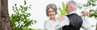 happy middle aged man in formal wear dancing with bride in white wedding dress in green garden, banner