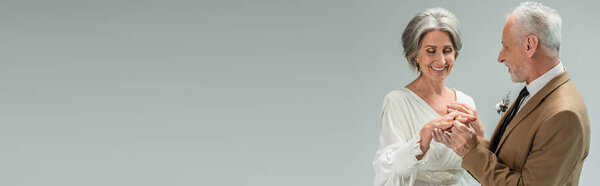 middle aged groom wearing golden engagement ring on happy bride in white dress isolated on grey, banner