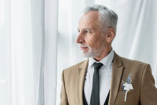 bearded middle aged man in suit with boutonniere looking away near white curtains