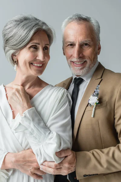 happy middle aged man in suit with floral boutonniere and woman in white wedding dress isolated on grey