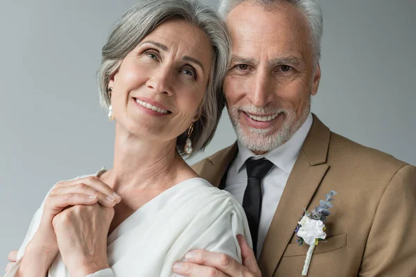 portrait of happy middle aged man in suit with floral boutonniere hugging woman in white wedding dress isolated on grey