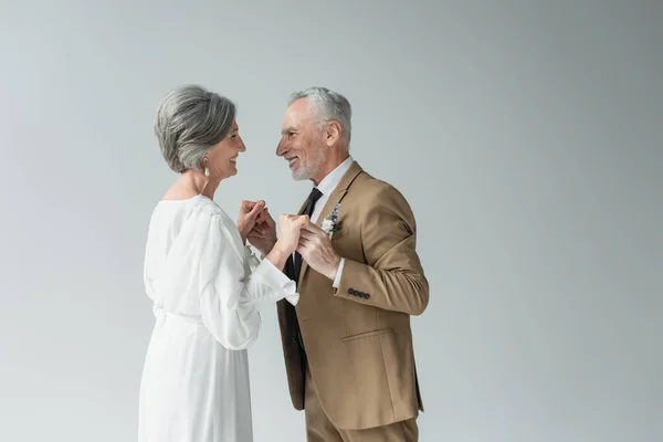 cheerful middle aged man in suit and bride in white wedding dress holding hands isolated on grey