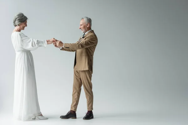 full length of cheerful middle aged man in suit and bride in white wedding dress holding hands on grey