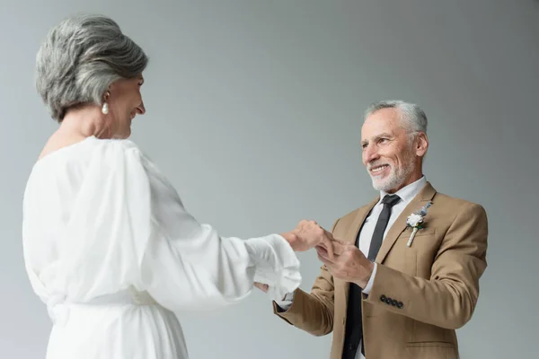 pleased middle aged man in suit with floral boutonniere and woman in white wedding dress holding hands isolated on grey