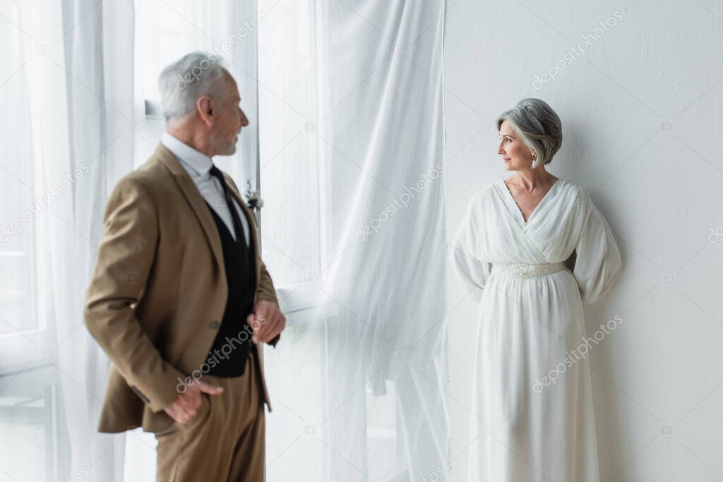 bearded middle aged man in suit posing while looking at cheerful bride in white dress near white curtains 