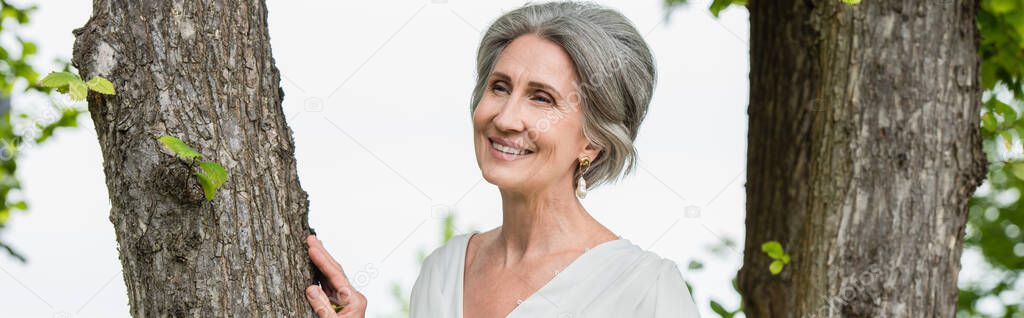 joyful middle aged woman in white dress touching tree trunk in park, banner
