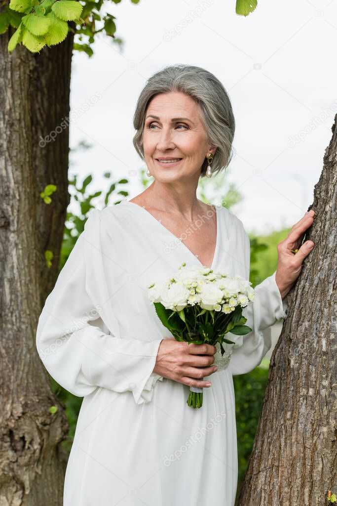 pleased middle aged bride in white dress holding wedding bouquet near tree trunk in park 