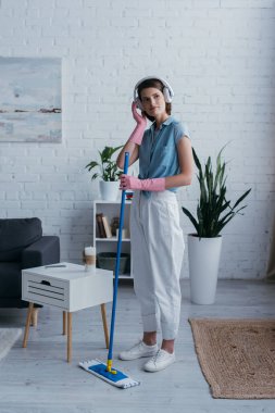 young woman adjusting wireless headphones while standing with mop in living room