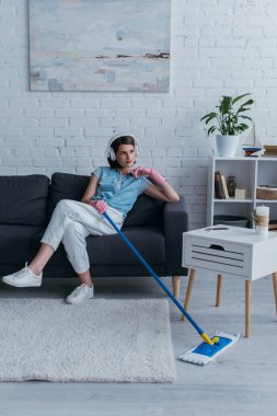 woman holding mop while sitting on couch and listening music in wireless headphones
