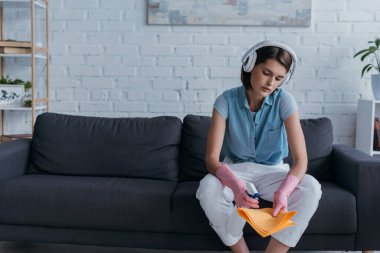woman in rubber gloves sitting on sofa in wireless headphones and spraying detergent on rag