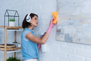 brunette woman in wireless headphones cleaning picture with rag and detergent