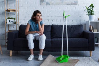 tired and thoughtful woman sitting on couch near broom and scoop