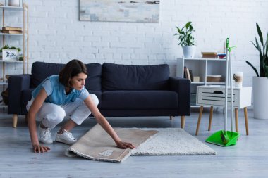 young woman moving carpet near broom and scoop in living room