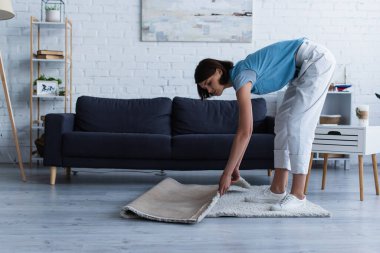 side view of young woman moving floor carpet in living room during house cleaning