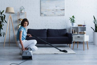 Brunette woman switching vacuum cleaner near carpet in living room 