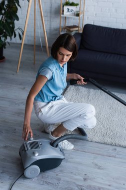 Brunette woman switching on vacuum cleaner in living room 