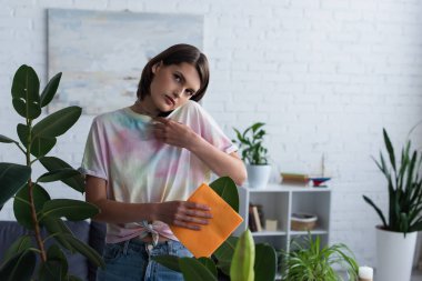 Young woman talking on smartphone while cleaning leaves of plant at home 