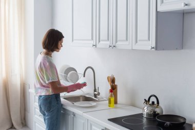 Side view of woman cleaning plate near dishwashing liquid in kitchen 