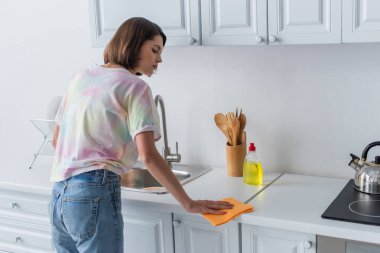 Side view of brunette woman cleaning worktop near sink in kitchen 