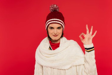 Smiling woman in knitted hat and scarf showing ok gesture isolated on red 