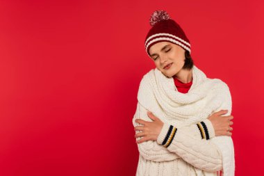 Pleased woman in knitted clothes closing eyes isolated on red 