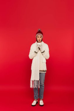 Full length of stylish woman in knitted clothes cooling drink in cup on red background