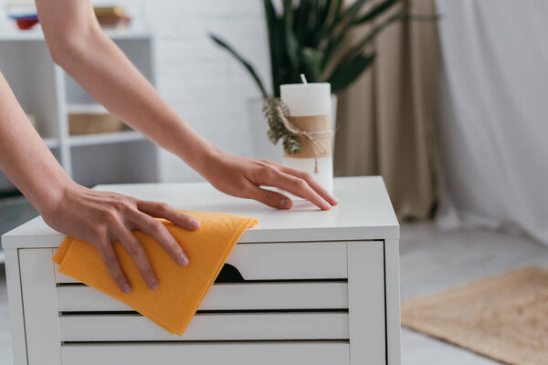 обрезанный вид молодой женщины с тряпкой вытирая белый прикроватный столик возле размытой свечи