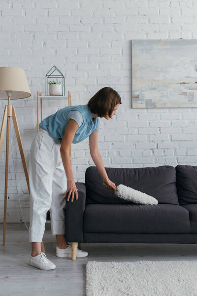 side view of brunette woman cleaning couch in living room at home