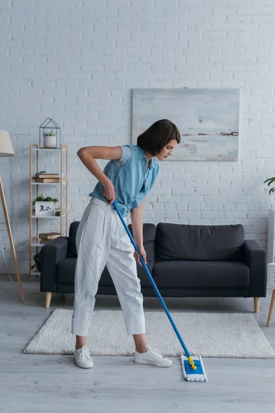 full length of young woman cleaning floor with mop near carpet