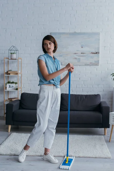 full length of young woman standing with mop in living room and looking away