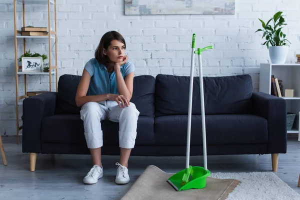 tired and thoughtful woman sitting on couch near broom and scoop
