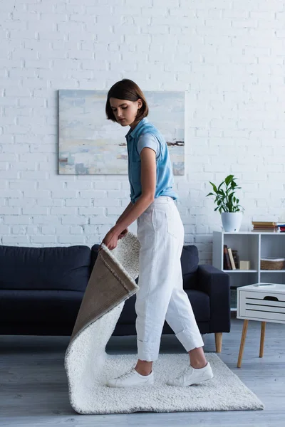 Young woman taking carpet from floor in living room 