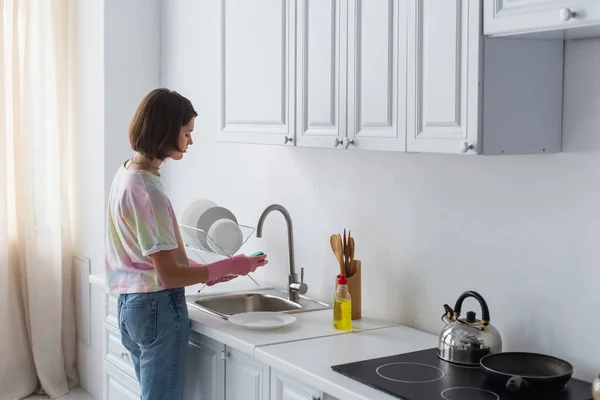 Side view of woman cleaning plate near dishwashing liquid in kitchen