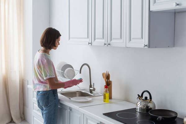 Side view of woman cleaning plate near dishwashing liquid in kitchen 