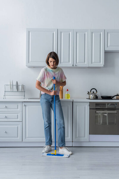 Brunette woman holding mop while cleaning floor in kitchen 