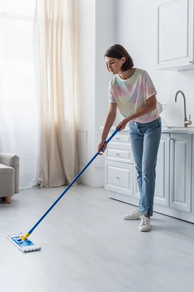 Brunette woman in casual clothes cleaning floor with mop in kitchen