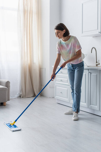 Brunette woman in casual clothes cleaning floor with mop in kitchen 