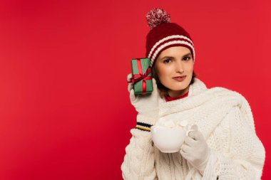 Smiling woman in hat and gloves holding gift near ear and cup with marshmallows isolated on red 