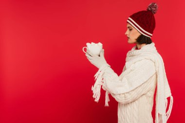 Side view of woman in warm clothes holding cup with marshmallows isolated on red 